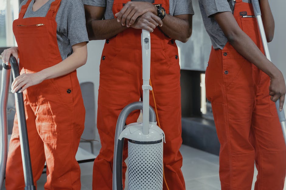 A person dressed in high-visibility orange workwear and a matching helmet, standing on a paved street near a concrete curb. The individual is facing away from the camera, observing a pile of mixed gar